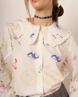 Camisa de popelina de algodón color crema con estampado de cerezas, dados, constelaciones y soles. Tiene cuello bobo con volantes y cierre frontal con botones.