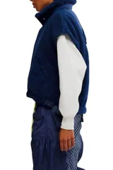 Chaleco de piel sintética tipo sherpa en color blanco crema, con cuello alto y cierre frontal mediante botones a presión. Presenta un diseño de hombros caídos y detalles de lazos decorativos en los laterales inferiores.