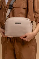 Bandolera compacta de textura símil cuero granulado color beige claro, con forma redondeada y cierre superior. Posee una placa metálica con el logo "LINCOLN'S" en el frente. Incluye una correa ajustable de tela negra con logo tejido en blanco.