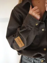 Camisa marrón de lana con corte clásico, cuello camisero, cierre frontal con botones de guampa, bolsillo de parche en el pecho izquierdo con logo bordado y puños abotonados.