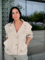 Chaqueta color nude con cuello solapa, cierre frontal de botones dorados y bolsillos laterales. Presenta detalles de pliegues horizontales en los hombros y verticales en la cintura.