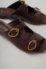 Sandalias planas de cuero color marrón con dos tiras anchas sobre el empeine, cada una con hebilla dorada.