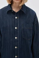 Campera tipo camisa de jean azul con rayas verticales blancas, corte holgado, cierre frontal con botones blancos y bolsillo de parche en el pecho.