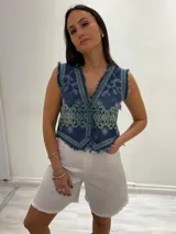 Chaleco corto de denim azul con bordados florales en blanco y marrón, detalle desflecado en escote, sisas y ruedo. Escote en V y cierre frontal con botones. Estilo boho chic.