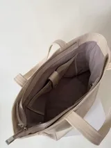 Cartera tipo tote color beige, hecha en cuero graneado con textura uniforme y suave al tacto. Tiene doble asa de hombro, un bolsillo frontal con broche imantado, un bolsillo trasero con cierre metálico y cierre principal también metálico. El interior está forrado y cuenta con tres bolsillos: uno con cierre, uno tipo plaqué para celular y un bolsillo grande tipo sobre con solapa y botón.