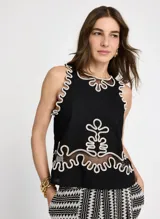 Blusa negra sin mangas con cuello redondo y detalles bordados en contraste color crudo en cuello, sisas y bajo. Presenta apliques ornamentales en el centro de la parte delantera y trasera, detalles calados con transparencias y abertura en forma de lágrima en la espalda con cierre de un botón.