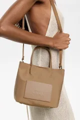 Bolso tote pequeño color marrón claro, de la marca Bimba y Lola, modelo Chihuahua. Tiene doble asa corta y correa larga extraíble. Presenta un bolsillo externo con parche con el logo de la marca.