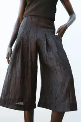 Pantalón culotte color marrón oscuro, de tiro alto con pliegues delanteros y tejido semitransparente efecto arrugado. Tiene forro interior y cierre lateral con cremallera oculta.