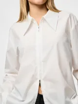 Camisa blanca de corte holgado, con cuello clásico, mangas largas con puños abotonados y cierre frontal con cremallera.