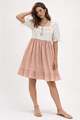 Vestido de dos tonos, con corpiño blanco de manga corta abullonada y escote cuadrado con bordado floral en tonos tierra. La falda es rosada, con frunce a la cintura y un ruedo con volado.