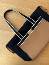 Bolso tipo tote de neopreno color negro y marrón, con cierre superior, bolsillo frontal marrón con logo rojo, bolsillo interno y externo con cierre, doble asa de mano marrón con detalles blancos y opción de correa larga.