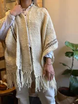 Ruana color crudo con rayas marrones tejidas en los bordes y flecos en el ruedo.