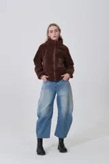 Campera de poliéster tipo sherpa en color natural, con diseño holgado, cuello alto y cierre frontal completo. Presenta un bolsillo de parche con cierre en el pecho y mangas largas con puños elásticos.