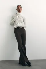 Pantalón de sarga color marrón oscuro, de tiro alto con elástico oculto en la cintura, cierre y pretina prolongada con botón y corchetes ocultos. Tiene piernas con corte para bota, bolsillos al bies, bolsillos de atrás ribeteados y pliegues adelante y atrás.