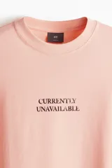 Remera color salmón de corte holgado, cuello redondo y manga corta. Estampado frontal con la frase "Currently Unavailable".