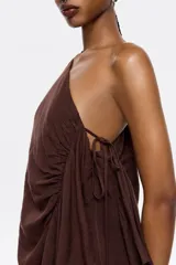Vestido largo marrón de lino, con corte ajustado y fruncido. Presenta escote de un solo hombro con lazo y maxitira de tejido. Cierre con cremallera invisible lateral y pañuelos laterales.