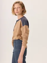 Buzo de manga larga con diseño de bloques de color y textura. El cuerpo principal es marrón, la espalda superior y hombros tienen un panel de tejido a rayas grises y azules, y los hombros superiores son de color beige rosado. Presenta costuras decorativas en rojo que separan los bloques de color.