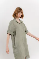 Vestido camisero corto color verde oliva, confeccionado en lino 100%. Tiene botones en la parte delantera, cuello camisero, mangas cortas con botón para arremangar y bolsillos laterales.