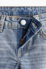 Jeans de denim azul claro con efecto lavado y desgaste sutil. Tienen un corte ajustado hasta la rodilla y luego se abren en un estilo acampanado (flare). Son de tiro bajo y presentan bolsillos delanteros abiertos y bolsillos traseros con solapa y botón.