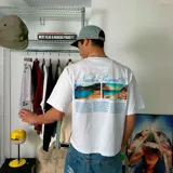 Camiseta blanca de corte holgado con estampado de fotos de playas y texto de la marca Nude Project en color celeste.