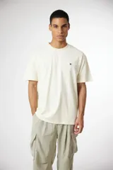 Remera de algodón color crema, corte oversize, con cuello redondo y un pequeño logo bordado en forma de 'x' en color azul marino en el pecho.