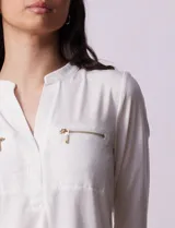 Blusa blanca de poliéster y elastano, con cuello a la base, escote en V y mangas tres cuartos. Presenta dos bolsillos frontales con cierre metálico dorado.