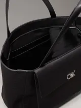 Cartera tipo shopper mediana de Calvin Klein, color negro, confeccionada en material textil con textura de lino tejido. Presenta asas fijas para el hombro, cierre de cremallera superior, bolsillo exterior frontal con cierre giratorio y herraje con monograma CK.