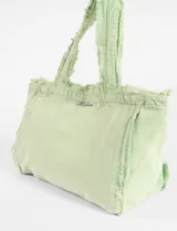 Bolso tipo tote de lona color verde claro, con efecto lavado o desgastado y bordes desflecados en las asas y la abertura superior. Presenta un bordado pequeño en el centro frontal.