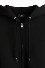 Campera con capucha y cierre frontal, color negro, confeccionada en tejido pesado de mezcla de algodón con interior cepillado suave. Presenta hombros caídos, capucha forrada con cordón ajustable y bolsillos delanteros tipo canguro. Puños y bajo acanalados.