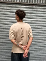 Remera oversize color beige con cuello redondo y manga corta. Presenta un pequeño corte o rasgadura en la parte delantera izquierda y estampados minimalistas en color marrón oscuro: la palabra "OSTEN" y un símbolo gráfico.