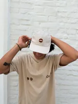 Remera oversize color beige con cuello redondo y manga corta. Presenta un estampado minimalista en color marrón en el pecho, que incluye un símbolo similar a un sol o estrella y texto.