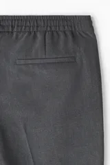 Pantalón jogger de vestir gris oscuro, corte slim fit, con cintura elástica ajustable con cordón, bolsillos laterales y traseros. Presenta pliegues delanteros y traseros.
