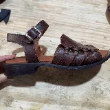 Sandalias bajas de cuero marrón con tiras anchas trenzadas en el empeine, tira con hebilla al tobillo y suela plana.