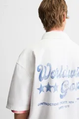 Camisa blanca de algodón con estructura canvas, corte boxy fit, cuello bowling y manga corta. Presenta bordado en azul claro con texto cursivo en el pecho y botones frontales.