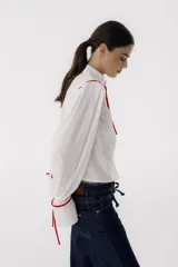 Camisa blanca de corte cropped con cuello mao y mangas largas abullonadas con lazos rojos en los puños. Presenta detalles de ribetes rojos en los hombros y lazos rojos en el cuello.