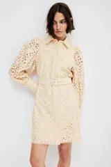 Vestido corto color beige con broderie, cuello camisero, botonadura frontal, cinturón extraíble con hebilla redonda, mangas globo largas con puños abotonados y ribetes festoneados.