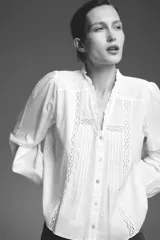 Camisa blanca de cuello subido y manga larga, con detalle de entredós combinados a tono y cierre frontal con botones.