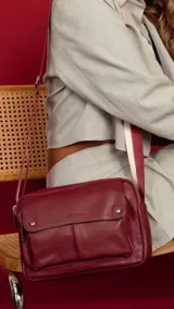 Bandolera bordeaux de cuero sintético con apertura central con cierre metálico. Tiene un bolsillo principal con cierre y un bolsillo frontal con tapa y broches magnéticos. Incluye bolsillos internos y posterior, y correa de galón combinada larga y regulable.