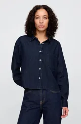 Camisa de jean corta color azul oscuro, con cuello clásico, cierre frontal con botones y bolsillo en el pecho.