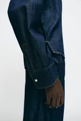 Camisa de denim azul oscuro, corte amplio, cuello solapa, manga larga y cierre frontal con botones a presión.