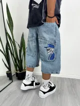 Bermuda de jean celeste de corte ancho y largo, estilo cargo, con múltiples bolsillos. Presenta un bordado de una calavera con gorra y cigarrillo en el bolsillo lateral derecho.