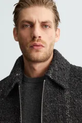 Sobrecamisa negra con textura de tejido bouclé y rayas verticales plateadas. Tiene cuello solapa, manga larga con puños abotonados, bolsillos laterales y cierre frontal con cremallera.