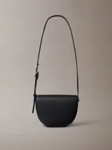 Cartera bandolera negra de cuero sintético con textura granulada, forma de media luna y correa fina ajustable. Cierre con solapa magnética y logo de Calvin Klein grabado en el frente.