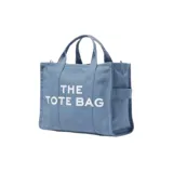 Bolso tote negro de lona con asas de mano y correa bandolera ajustable. Estampado frontal con la leyenda "THE TOTE BAG MARC JACOBS" en letras blancas.