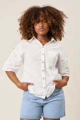 Camisa color beige claro, de corte clásico, con cuello, botones frontales, bolsillo en el pecho y mangas tres cuartos con presilla para sujetar.