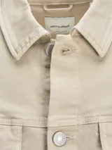 Campera de denim color beige, de corte recto, con cuello clásico, cierre frontal con botones metálicos, dos bolsillos con solapa abotonada en el pecho y puños abotonados.