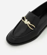 Zapato mocasín femenino color marrón oscuro, confeccionado en poliuretano, con detalle metálico dorado en forma de lazo en el empeine y tacón bajo.
