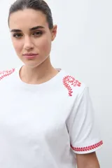 Remera blanca de algodón con cuello redondo y mangas cortas. Presenta bordados florales rojos en los hombros y en los bordes de las mangas.