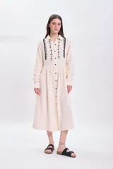 Vestido largo de muselina color crudo con bordado negro en el cuello y la pechera, corte evasé, manga larga y abotonadura frontal.