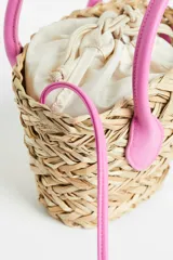 Cartera bandolera de paja color beige, con forro de tela color crema con cierre de cordón ajustable. Tiene dos asas cortas y correa larga ajustable de color rosa.